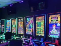 Orange County encerra operação ilegal de jogos de azar disfarçada de negócio de portas de garagem Orange County encerra operação ilegal de jogos de azar disfarçada de negócio de portas de garagem. Uma fileira de máquinas de jogos estilo caça-níqueis bem iluminadas com telas digitais coloridas fica dentro de uma sala escura, cada máquina brilhando com contornos de néon e exibindo vários gráficos de jogo. Cadeiras vazias e garrafas de bebidas são posicionadas em frente às máquinas.