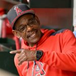 Ron Washington com um moletom vermelho e boné do Los Angeles Angels, sorrindo e apontando para o banco de reservas.