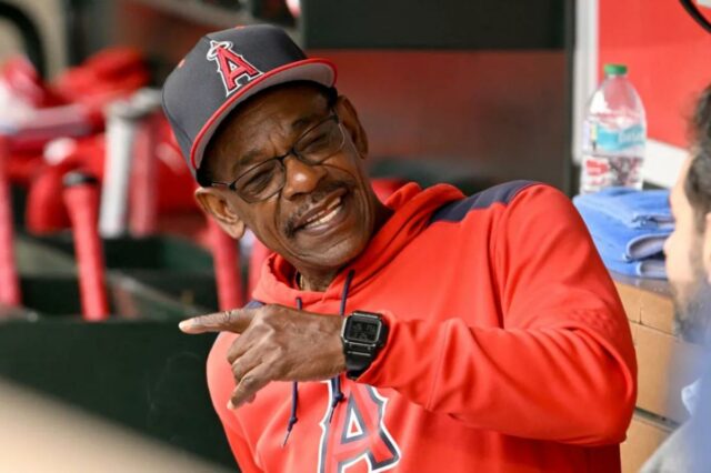 Ron Washington com um moletom vermelho e boné do Los Angeles Angels, sorrindo e apontando para o banco de reservas.