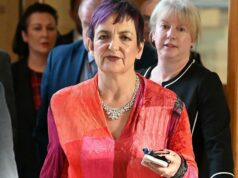 Secretário de Educação rejeita chamadas de investigação de Constance Douglas Ross, de terno escuro, camisa azul e gravata cor de vinho, está de pé no parlamento. Ele está gesticulando com as duas mãos, claramente defendendo um ponto de vista em um debate.