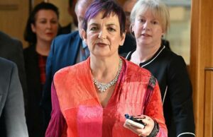 Secretário de Educação rejeita chamadas de investigação de Constance Douglas Ross, de terno escuro, camisa azul e gravata cor de vinho, está de pé no parlamento. Ele está gesticulando com as duas mãos, claramente defendendo um ponto de vista em um debate.