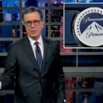 Stephen Colbert de terno gesticulando com a mão esquerda em frente ao horizonte da cidade.