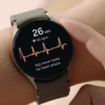 Samsung Galaxy Watch 7 em um suporte, mostrando a tela inicial com outros produtos Galaxy em segundo plano