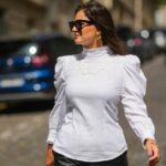 Amanda Mühle é vista vestindo um casaco de lã cinza com lapelas largas, fecho de botão e gola abotoada da Siyazu; um suéter de malha estampado em tom suave de verde e rosa com padrão argyle e textura de mistura de lã macia da Marc O'Polo Denim; uma camiseta branca de algodão da COS usada por baixo; jeans azul com corte reto e solto da Zara; botas Mou de camurça bege com sola plataforma e detalhes costurados exclusivos da Mou; uma “bolsa City” Balenciaga de couro rosa com formato suavemente desleixado, alças superiores e detalhes prateados da Balenciaga; vários anéis de prata da Pilgrim; segurando uma revista Vogue; óculos estilo aviador com armação rosa e lentes levemente coloridas da Lexxola; brincos de prata com acabamento escultural da Mango; seus longos cabelos castanhos estão soltos e repartidos de lado em 8 de janeiro de 2026 em Munique, Alemanha.