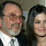 Uma mulher loira está rindo e segurando um cachorrinho vestido com uma roupa roxa e rosa. Ela está vestida com uma jaqueta rosa brilhante, óculos escuros vermelhos e um telefone rosa preso à orelha. O fundo mostra um ambiente externo desfocado com pessoas e vegetação.