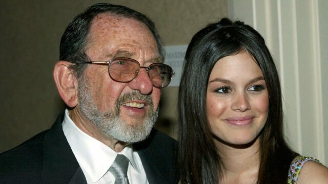 Uma mulher loira está rindo e segurando um cachorrinho vestido com uma roupa roxa e rosa. Ela está vestida com uma jaqueta rosa brilhante, óculos escuros vermelhos e um telefone rosa preso à orelha. O fundo mostra um ambiente externo desfocado com pessoas e vegetação.