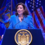 Discurso sobre o estado do estado do governador Hochul, ela está vestindo uma camisa azul e está no palco em frente a um pódio. A Comissão de Jogos de Nova York aplaude Hochul porque o estado tem como alvo as apostas online para menores de idade