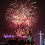 Um homem com pêlos faciais curtos e brancos olha para a câmera. Ele usa um chapéu preto, óculos pretos e uma capa de chuva preta. Ele está do lado de fora, no escuro, em frente a um palco bem iluminado