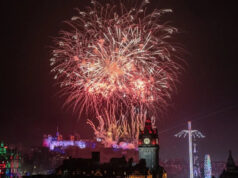 A Escócia comemora o ano novo com o início do alerta meteorológico em 2026 Um homem com pêlos faciais curtos e brancos olha para a câmera. Ele usa um chapéu preto, óculos pretos e uma capa de chuva preta. Ele está do lado de fora, no escuro, em frente a um palco bem iluminado