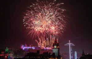 A Escócia comemora o ano novo com o início do alerta meteorológico em 2026 Um homem com pêlos faciais curtos e brancos olha para a câmera. Ele usa um chapéu preto, óculos pretos e uma capa de chuva preta. Ele está do lado de fora, no escuro, em frente a um palco bem iluminado