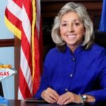 Dina Titus sentada à sua mesa em um escritório do governo, sorrindo em uma jaqueta azul, com bandeiras dos EUA e de Nevada atrás dela e uma placa “Bem-vindo à Fabulosa Las Vegas” na mesa. A deputada Dina Titus insta o Congresso a restaurar as deduções totais do jogo para proteger os consumidores