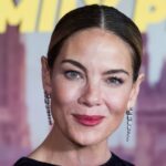 Uma mulher madura usa sua máscara facial de LED vermelho (terapia de luz vermelha) enquanto relaxa em casa, em uma poltrona. Ela usa seu smartphone - Fotografia de stock