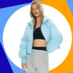 Uma pessoa vestindo uma jaqueta verde escura, gola alta preta, calça jeans e faixa preta.