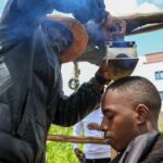 O barbeiro e criador de conteúdo viral Safari Martins demonstra um de seus métodos inventivos de barbear usando uma caixa de ferro enquanto prepara Ian Njenga em Kiambu, Quênia, quarta-feira, 26 de novembro de 2025. (AP Photo / Andrew Kasuku)