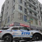 Um carro da polícia de Nova York estacionado em uma rua coberta de neve ao lado de um prédio com uma farmácia no térreo.