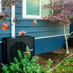 A dark grey heat pump sits outside a blue home.