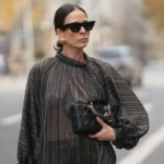 Amanda Mühle é vista vestindo um casaco de lã cinza com lapelas largas, fecho de botão e gola abotoada da Siyazu; um suéter de malha estampado em tom suave de verde e rosa com padrão argyle e textura de mistura de lã macia da Marc O'Polo Denim; uma camiseta branca de algodão da COS usada por baixo; jeans azul com corte reto e solto da Zara; botas Mou de camurça bege com sola plataforma e detalhes costurados exclusivos da Mou; uma “bolsa City” Balenciaga de couro rosa com formato suavemente desleixado, alças superiores e detalhes prateados da Balenciaga; vários anéis de prata da Pilgrim; segurando uma revista Vogue; óculos estilo aviador com armação rosa e lentes levemente coloridas da Lexxola; brincos de prata com acabamento escultural da Mango; seus longos cabelos castanhos estão soltos e repartidos de lado em 8 de janeiro de 2026 em Munique, Alemanha.