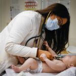 John Aguilar, de sete meses, é segurado por sua mãe Kimberly Fernandez, à esquerda, e pela Dra. Vidya Mony, à direita, enquanto recebe uma vacina de Farah Ng, RN, na clínica Valley Med em San Jose, Califórnia, na quinta-feira, 8 de janeiro de 2026. (Shae Hammond/Bay Area News Group)