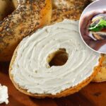 Bagel com sementes de papoula, torrado e barrado com cream cheese, com uma faca e uma tigela de cream cheese.