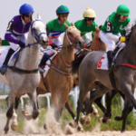 Entain anuncia que encerrará o patrocínio da Coral Cup após 52 anos. Um grupo de jóqueis em sedas coloridas de corrida montam cavalos galopando juntos durante uma corrida de cavalos, com poeira levantando da pista enquanto eles fazem uma curva.