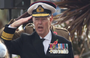 Espada suspeita e medalha rara – como o falso almirante foi finalmente descoberto Um homem vestido de almirante faz uma saudação. Ele usa chapéu branco, jaqueta azul marinho, camisa branca e gravata. No peito há uma série de medalhas penduradas por fitas coloridas.