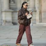 Patricia Wirschke é vista vestindo um clássico casaco longo Herno bege, com gola entalhada, aba de proteção e punhos com cinto; um conjunto Incentive cor bordô com um suéter tricotado, com bainha, punhos e decote canelados e calças tricotadas de perna larga com corte descontraído; um par de mocassins Saint Laurent de camurça macia Le Loafer em marrom escuro com detalhe de logotipo dourado na lateral; uma bolsa Hop marrom escuro da Bottega Veneta com a famosa construção em couro Intrecciato, tecida; Óculos de sol pretos com armação quadrada Tom Ford Ilias e lentes em tom laranja; Brincos dourados Bvlgari Serpenti Viper; seu cabelo castanho é penteado em um estilo liso, na altura dos ombros, com uma parte central; ela segura uma revista enrolada nas mãos em 19 de agosto de 2025 em Duesseldorf, Alemanha.