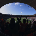 FIFA x Stats Perform. Torcedores comemorando dentro de um estádio de futebol lotado durante uma partida, vistos através de uma lente grande angular sob um teto oval aberto.