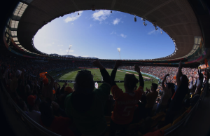 FIFA assina direitos exclusivos da Copa do Mundo de 2026 para a Stats Perform como distribuidora oficial global de direitos de apostas FIFA x Stats Perform. Torcedores comemorando dentro de um estádio de futebol lotado durante uma partida, vistos através de uma lente grande angular sob um teto oval aberto.