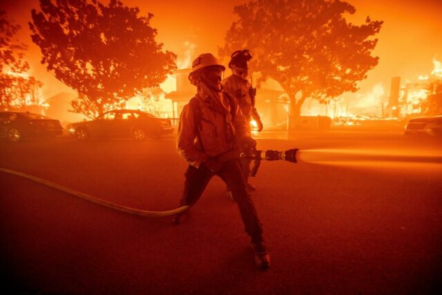 Dois bombeiros com equipamento de proteção borrifam água de uma mangueira enquanto chamas e fumaça engolfam as casas ao fundo.