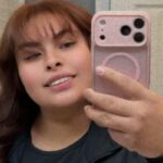 Bianca Esquivel, uma mulher de 24 anos com cabelos castanho-avermelhados e franja, sorri enquanto tira uma selfie com um celular rosa.