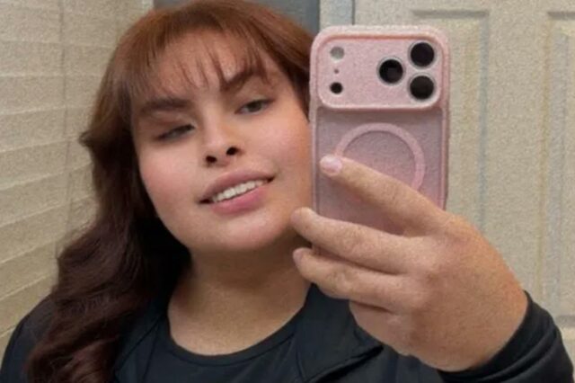 Bianca Esquivel, uma mulher de 24 anos com cabelos castanho-avermelhados e franja, sorri enquanto tira uma selfie com um celular rosa.