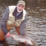 Um homem de cabelos brancos e óculos gesticula com a mão enquanto fala em frente a uma janela.