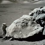 Quatro astronautas em trajes espaciais azuis estão na grama com um foguete laranja ao fundo.