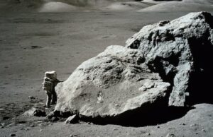 Já estivemos na lua, então por que vamos de novo? Quatro astronautas em trajes espaciais azuis estão na grama com um foguete laranja ao fundo.