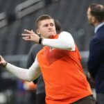 Bailey Zappe, do Cleveland Browns, se aquece, vestindo uma camisa branca de mangas compridas sob um colete laranja e segurando uma bola de futebol.