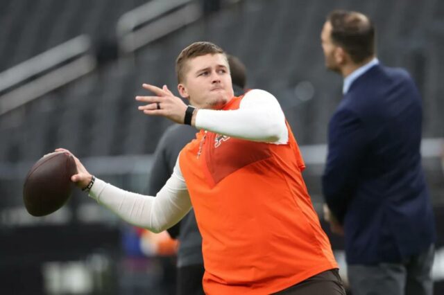 Bailey Zappe, do Cleveland Browns, se aquece, vestindo uma camisa branca de mangas compridas sob um colete laranja e segurando uma bola de futebol.