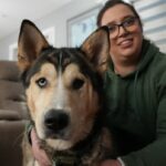 Mia Ribeck é uma mulher branca com uma touca preta da Hello Kitty com um pom pom no topo. Ela está vestindo uma parca preta e botas de borracha de cano alto. Ela está agachada ao lado de sua cadela Greta, uma grande Doberman preta.