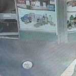 A imagem do CCTV mostra dois homens passando por uma pequena loja. Um homem usa chapéu preto, casaco preto, calça cinza e camisa pólo. Outro usa o que parece ser uma jaqueta vermelha e um chapéu escuro.