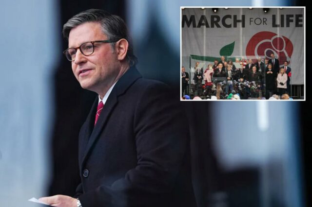 Um homem com cabelos grisalhos e óculos, vestindo um casaco preto e gravata vermelha, falando em um pódio.