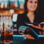 Um cliente pagando com um smartphone tocando em um terminal de pagamento segurado por um barman sorridente.