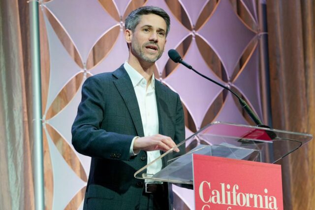 Um homem com cabelos grisalhos e barba falando em um pódio com 