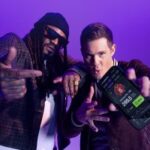 Marshawn Lynch e Adam Devine posam contra um fundo roxo, gesticulando em direção à câmera enquanto Adam Devine segura um smartphone exibindo o aplicativo de esportes de fantasia PrizePicks. PrizePicks se junta a Marshawn Lynch e Adam Devine para um anúncio de apostas irônico