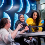 Uma foto de Imad Ahmed. Ele é careca, com bigode preto e cinza e cavanhaque. Ele está usando óculos e um lenço cinza.