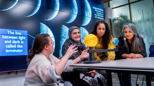 Uma foto de Imad Ahmed. Ele é careca, com bigode preto e cinza e cavanhaque. Ele está usando óculos e um lenço cinza.