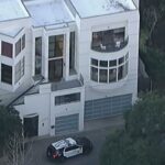 Vista aérea de uma casa branca de vários andares com um carro da polícia estacionado em frente.