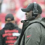 O técnico do Tampa Bay Buccaneers, Todd Bowles, usando um fone de ouvido e um boné preto com logotipo, assistindo da linha lateral.