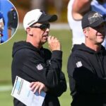 Dois treinadores de futebol assistindo a um jogo, um deles usando óculos escuros e segurando um papel.