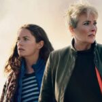 Two women stand in front of a cloudy sky looking in opposite directions