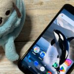 Uma mão segurando um smartphone Google Pixel azul com um ícone de alerta e ícones representando câmera, áudio e bugs.