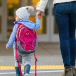 Vista traseira de uma jovem mãe alegre segurando a mão da filha enquanto caminhava na faixa de pedestres até a escola da criança.